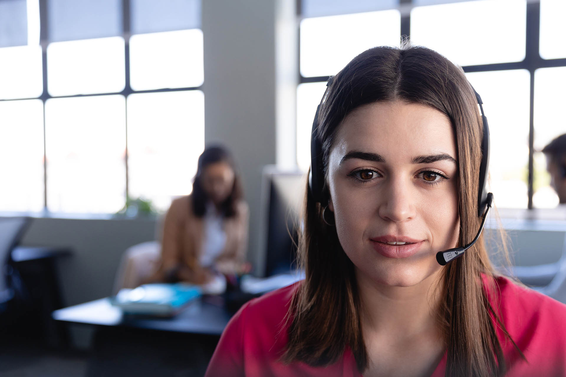 Front view close up of a mixed race woman working as a customer support, wearing smart clothes and headset, using a desktop computer with colleagues working in the background. customer support